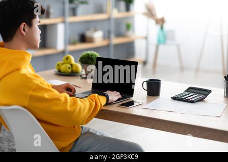 Asian Man Working On Laptop Computer With Blank Black Screen At Home Office, Sitting At Desk On Chair, Over Shoulder View, Mockup. People, Technology, Stock Photo