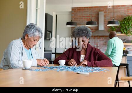 Two diverse female friends sitting in kitchen with coffee and doing puzzles Stock Photo