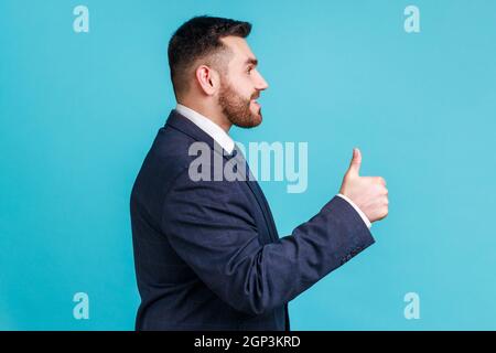 Well done, good job! Profile portrait of smiling happy bearded man wearing dark official style suit, looking ahead, showing thumb up. Indoor studio shot isolated on blue background. Stock Photo