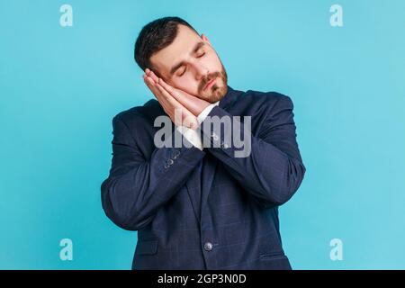 Bedtime. Portrait of handsome bearded man in suit sleeping laying down on her palms and smiling pleased, having comfortable nap and resting, dozing off. Indoor studio shot isolated on blue background. Stock Photo