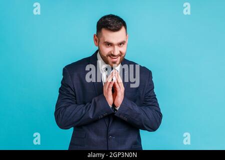 Portrait of devious handsome brunette man with beard wearing dark official style suit, thinking devious tricks and cheats, looking at camera. Indoor studio shot isolated on blue background. Stock Photo