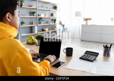 Over the shoulder view of busy asian guy working on laptop in home office, male looking at blank computer screen and typing on keyboard, filling repor Stock Photo