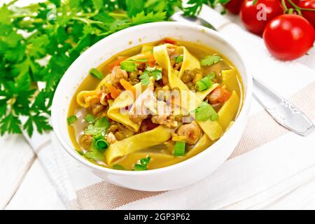 Soup with meat, tomatoes, vegetables, mung bean lentils and noodles in a bowl on a napkin, parsley and spoon on the background of light wooden board Stock Photo