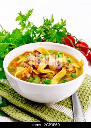 Soup with meat, tomatoes, vegetables, mung bean lentils and noodles in a bowl on a green napkin, parsley and spoon on the background of light wooden b Stock Photo