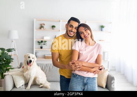 Portrait of happy multiracial couple hugging and smiling at camera, their adorable pet dog sitting on couch nearby, free space. Loving Arab guy embrac Stock Photo