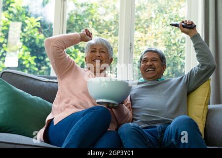 Two diverse senior couple sitting on sofa, watching tv and having fun Stock Photo