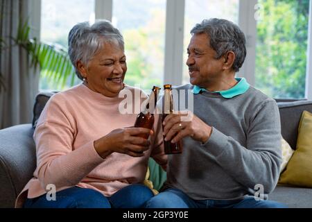 Happy diverse senior couple sitting on sofa and drinking beer Stock Photo