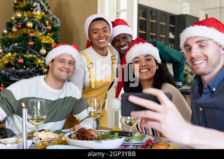 Group of happy diverse female and male friends in santa hats , celebrating christmas, taking selfie Stock Photo
