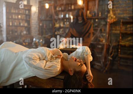 Male exorcist in black hood casting out demons from crazy woman. Exorcism, mystery paranormal ritual, dark religion, night horror, potions on shelf on Stock Photo