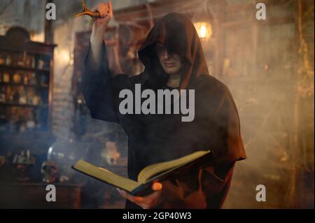 Male exorcist in black hood holds book of spells. Exorcism, mystery paranormal ritual, dark religion, night horror, potions on shelf on background Stock Photo