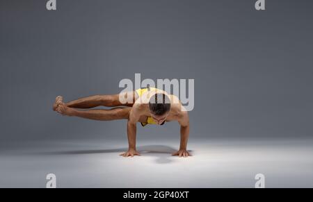Muscular yoga stands on hands in difficult pose, meditation position, grey background. Strong man doing yogi exercise, asana training Stock Photo