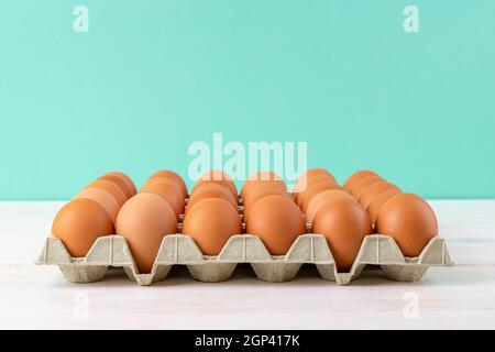 Brown chicken eggs in the cardboard tray on a white wooden table against turquoise background. Raw organic hen eggs with brown shell for Easter. Stock Photo