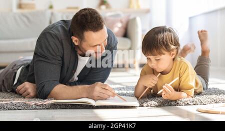 Kids development and art hobby concept. Young loving father drawing with his little son, painting in album with pencils, lying on floor at home, panor Stock Photo