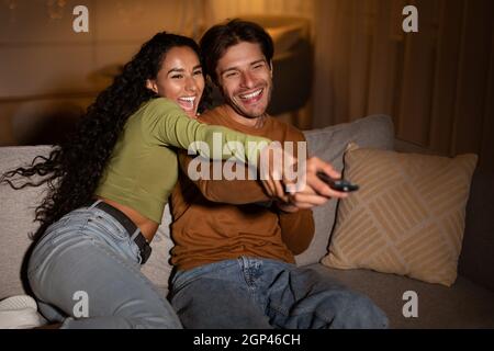 Joyful Couple Of Viewers Watching TV Pointing Remote Control And Switching Channels Having Fun Sitting At Home In The Evening. Weekend Fun And Family Stock Photo