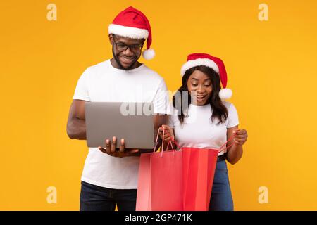 Black Couple Shopping Using Laptop During Christmas Sale Holding Colorful Paper Shopper Bags Over Yellow Studio Background. Happy Spouses In Santa Hat Stock Photo