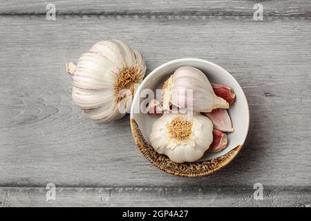 Table top view on garlic bulbs and peeled cloves in small ceramic bowl placed on gray wood desk. Stock Photo