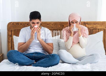 Muslim pregnant couple got flu, having runny nose, sitting on bed and blowing nose, sneezing in paper tissue, feeling unwell. Rhinitis and seasonal al Stock Photo