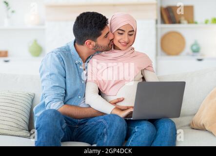 Muslim expecting woman and her loving husband using laptop, relaxing on sofa at home and searching furniture for baby room, copy space. Caring man kis Stock Photo
