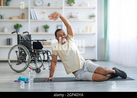 Yoga for paraplegic patients. Disabled black guy stretching on sports mat at home, wheelchair standing nearby. Cheerful impaired young man doing flexi Stock Photo