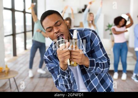 Attractive black guy with beer bottle singing karaoke on party with international friends, performing song into mic at home. Cool African American man Stock Photo