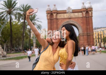 Smiling diverse young girlfriends embracing happily while taking selfie on smartphone against famous triumphal arch during summer tour together in Barcelona city Stock Photo