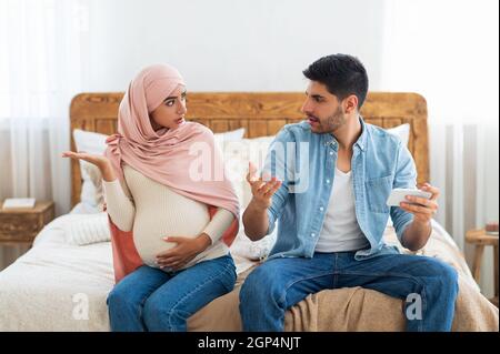 Pregnancy and relationship problems. Muslim couple arguing at home, expectant arab lady in hijab and her husband quarreling while sitting on bed, free Stock Photo