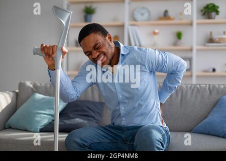 Young African American guy suffering from back pain, leaning on crutch, having difficulty getting up from sofa at home. Millennial black man having in Stock Photo