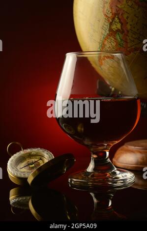 A shallow depth of field focus on a glass of Cognac with a compass and antique globe with a dark reflective red and black background. Stock Photo