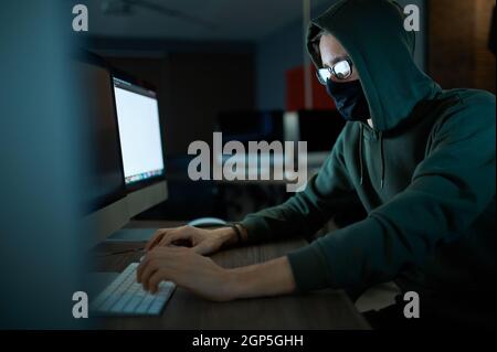 Male internet hacker in hood holds laptop, front view. Illegal web programmer at workplace, criminal occupation. Data hacking, cyber security Stock Photo