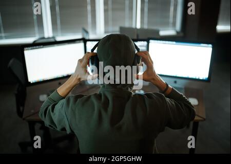 Male internet hacker in hood and headphones works on computer. Illegal web programmer at workplace, criminal occupation. Data hacking, cyber security Stock Photo