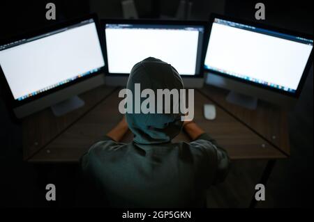Male internet hacker in hood sitting at monitors, back view. Illegal web programmer at workplace, criminal occupation. Data hacking, cyber security Stock Photo