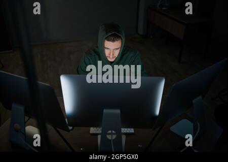 Male internet hacker in hood works on computer, front view. Illegal web programmer at workplace, criminal occupation. Data hacking, cyber security Stock Photo