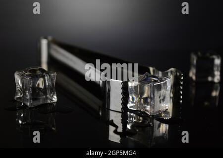 A pair of tongs next to some fresh ice cubes over a dark reflective surface. Stock Photo