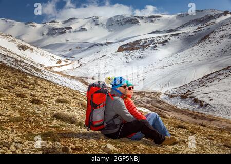 Happy Young Couple Sitting and Hugging in the Mountains at the Base Camp. Enjoying beautiful View on Snowy Mountains. Active Sportive Lifestyle. Winte Stock Photo