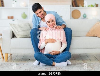 Caring young arab man massaging his wife's shoulders, resting together at home in living room interior. Happy muslim man taking care of his expectant Stock Photo