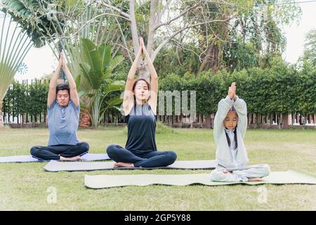 Asian young mother, father practicing doing yoga exercises with child daughter outdoors in meditate pose together in nature a field garden park, famil Stock Photo