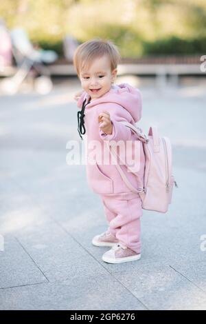 Cute funny girl 1-2 year old wear pink sweatshirt with hood and backpack walking in city street over urban outdoors. Childhood. Little toddler child m Stock Photo