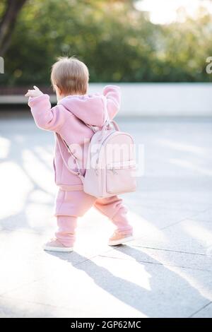 Funny baby girl 1 year old wear stylish pink sport suit and backpack walk on street over urban background. Cute little child with trendy clothes. Chil Stock Photo