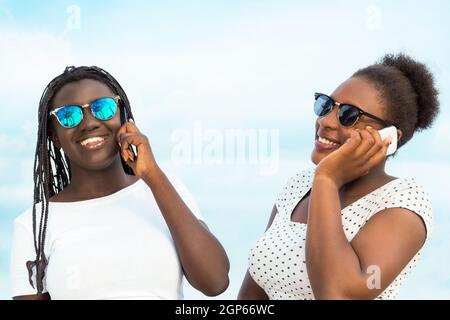 Close up portrait of two diverse african teen girls wearing sun glasses talking on smart phones outdoors. Stock Photo