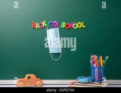 inscription back to school from multi-colored plastic letter on a green chalk board, hanging disposable medical mask Stock Photo