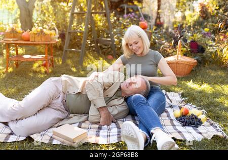 Tender senior couple enjoying time together, resting on picnic blanket in garden and chatting, man lying on wife's legs. Happy retirement concept Stock Photo