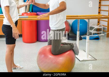 Man kneeling on a stability ball, doing exercises for a strong core with the assistance of a female therapist, at the rehabilitation center Stock Photo