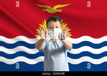 Little white boy in a protective mask on the background of the flag of Kiribati Makes a stop sign with his hands, stay at home Kiribati. Stock Photo