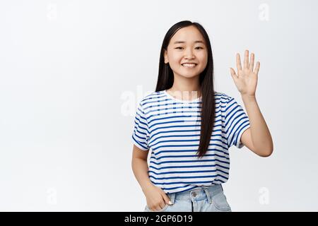 Friendly asian teen girl saying hi, waving raised hand and smiling happy at camera, greeting you, standing over white background Stock Photo