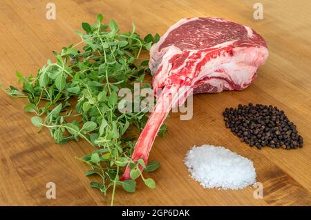 Raw angus Tomahawk steak on a wooden board with salt, pepper, oregano and a knife Stock Photo