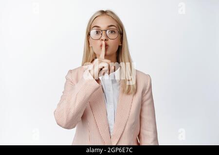 Cute business woman in glasses and suit shushing, making hush sign with finger on lips, keeping secret, standing over white background Stock Photo