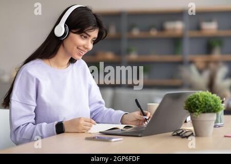 Portrait of happy woman in headset taking notes working on laptop computer at home office, free space. Freelancer noting down information from interne Stock Photo