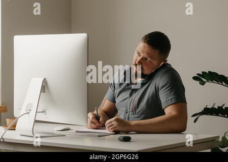 A man works remotely on a desktop computer. A guy with a beard is taking notes during a phone calling and staring at the display of the computer at ho Stock Photo