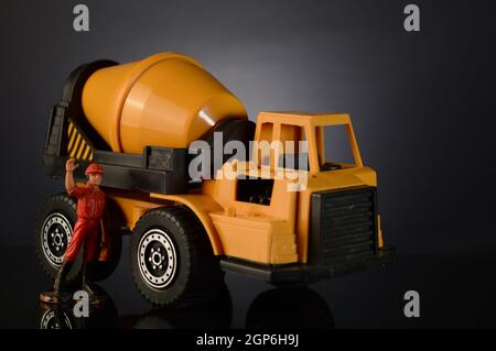A toy cement mixer and a miniature working man waving over a dark gradient background. Stock Photo