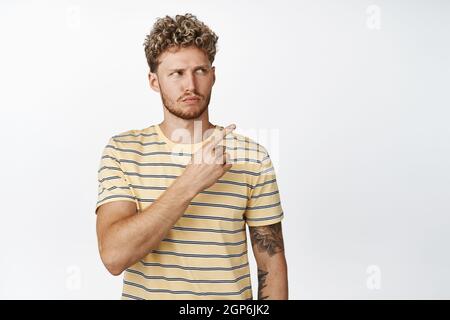 Disappointed young blond guy, looks skeptical at empty space for brand name or product, points finger at advertisement with doubtful expression, white Stock Photo